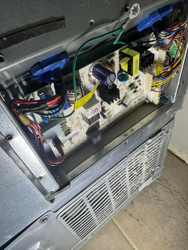 Appliance main control board with relays and a transformer, exposed behind its service panel with wiring harnesses connected