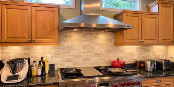 Stainless steel chimney range hood with its lights on, installed above a Wolf range with red knobs and a center griddle, between oak cabinets