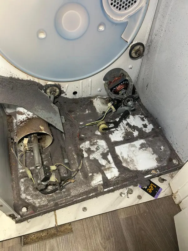 Gas dryer with the drum removed, lint covering the base around the burner assembly, drive motor and idler pulley, with coins on the floor of the cabinet