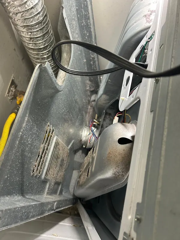 Inside a gas dryer cabinet: lint on the base and burner shield, the drive belt off, and a yellow gas supply line behind