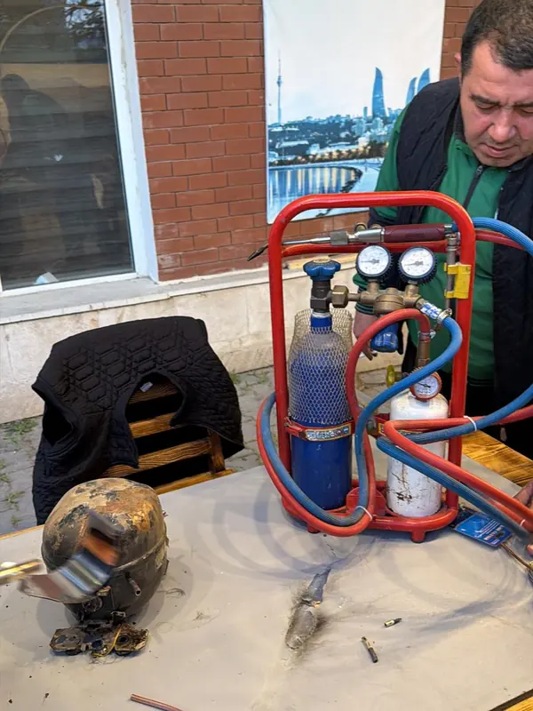 Technician brazing copper refrigerant lines beside a removed refrigerator compressor, with an oxygen and fuel torch kit on the bench