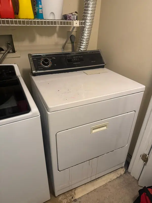 Older white Whirlpool heavy-duty dryer with a flexible vent duct running up the wall behind it